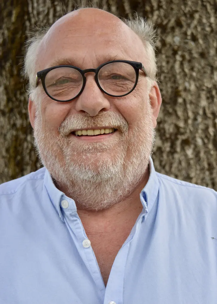 En leende man med grått hår på sidorna av huvudet. Kal på hjässan. Han har kortväxt skägg och ett par svarta runda glasögon. Hans ljusblå skjorta är lite uppknäppt vid halsen. Bilden är tagen utomhus framför en trädstam.