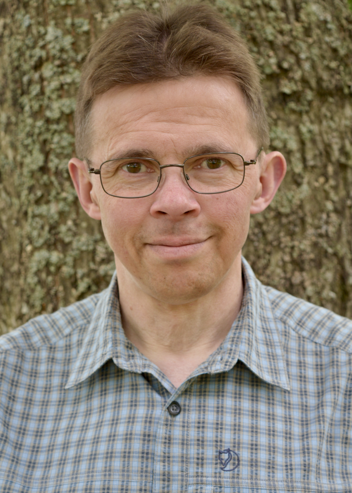 En leende man med ljusbrunt kortklippt hår. Han har fyrkantiga glasögon på sig. Skjortan han har på sig är rutig, ljusblå och grå. Han står utomhus framför en trädstam.