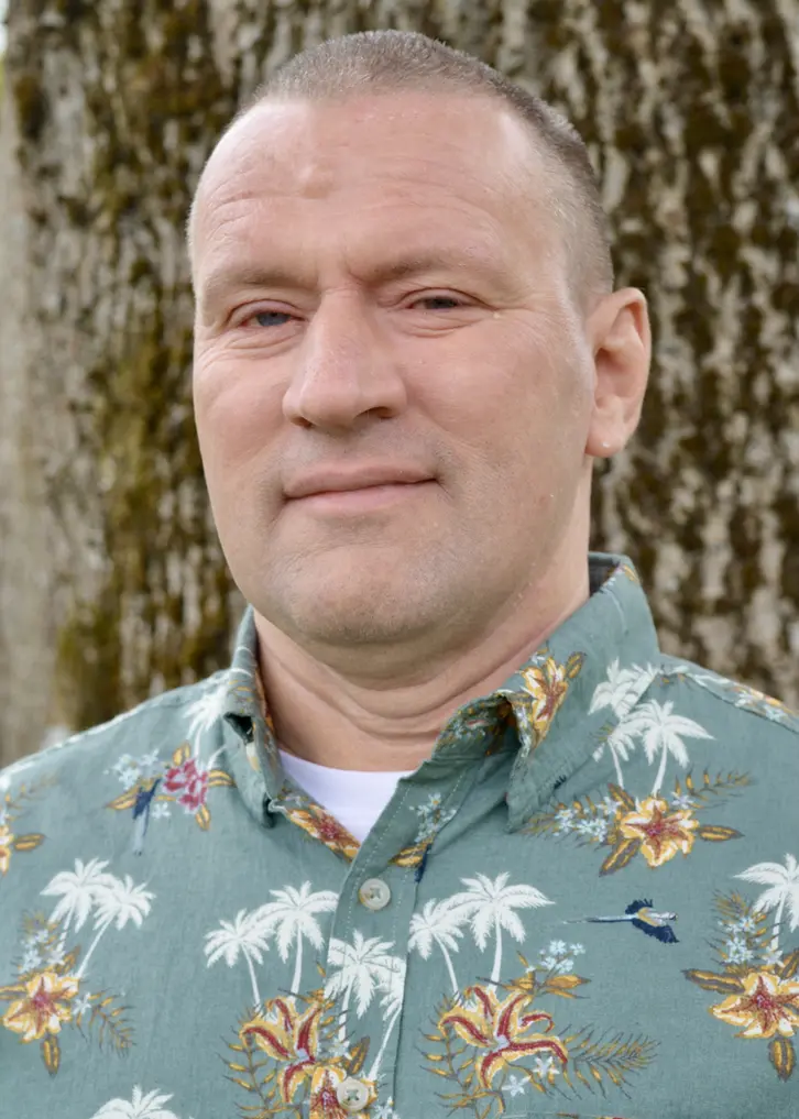 En man med brunt hår som är mycket kortklippt. Han har en grågrön skjorta på sig med vita palmer och blommor i orange. Under skjortan syns en vit T-shirt. Bakom honom syns en trädstam med lite mossa. Bilden är tagen utomhus.