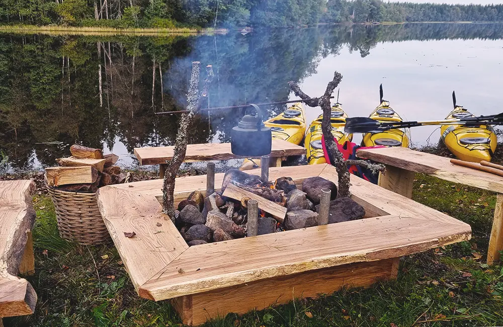 Grillplats med eldrök. I bakgrunden hägrar en spegelblank sjö och på stranden finns fyra kajaker parkerade.