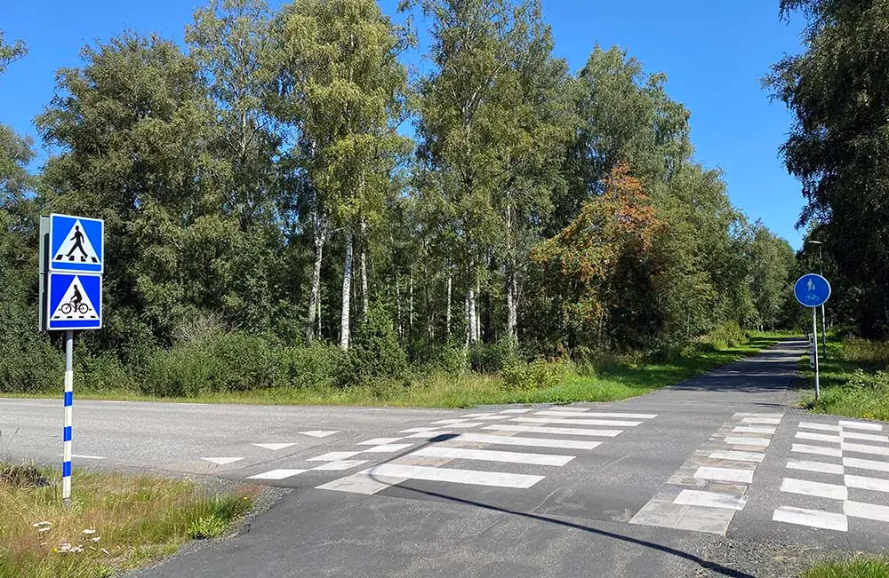 En cykelöverfart med övergångsställe, vägbula och vägskyltar som visar att cyklister och gångtrafikanter har företräde. I bakgrunden syns lummiga gröna träd och blå himmel.