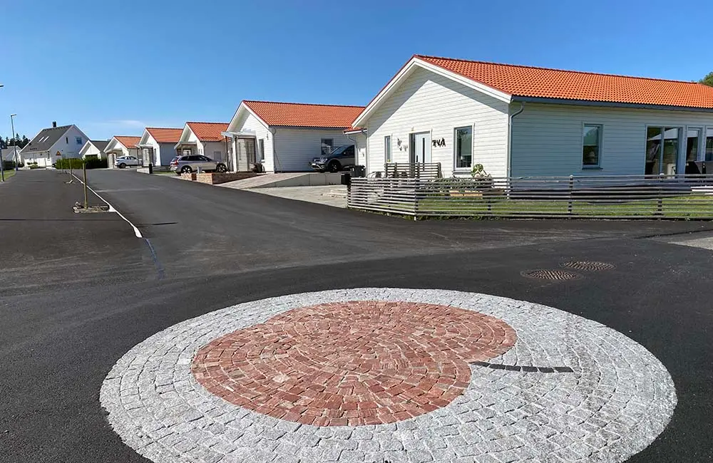 Ett nybyggt bostadsområde med likadana vita enplansvillor. Himlen är blå och taken är tegelfärgade. Första huset har e grön gräsmatta. I förgrunden finns en rund stenläggning som föreställer ett äpple.