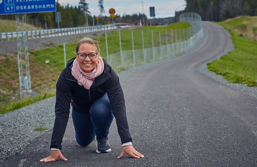 Kvinna med svart jacka och rosa halsduk står i startposition på en cykelbana längst med en motorväg. Vid sidan av vägen är det gräs.