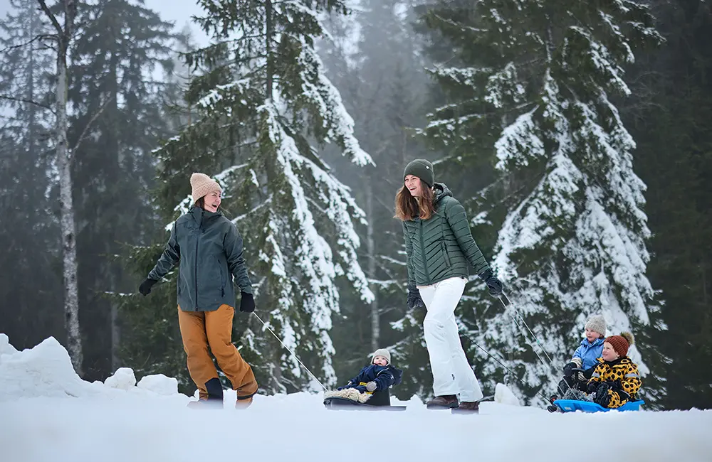 Två kvinnor i vinterkläder drar tre barn i pulka bakom sig på snön. I bakgrunden är träden täckta i snö. 