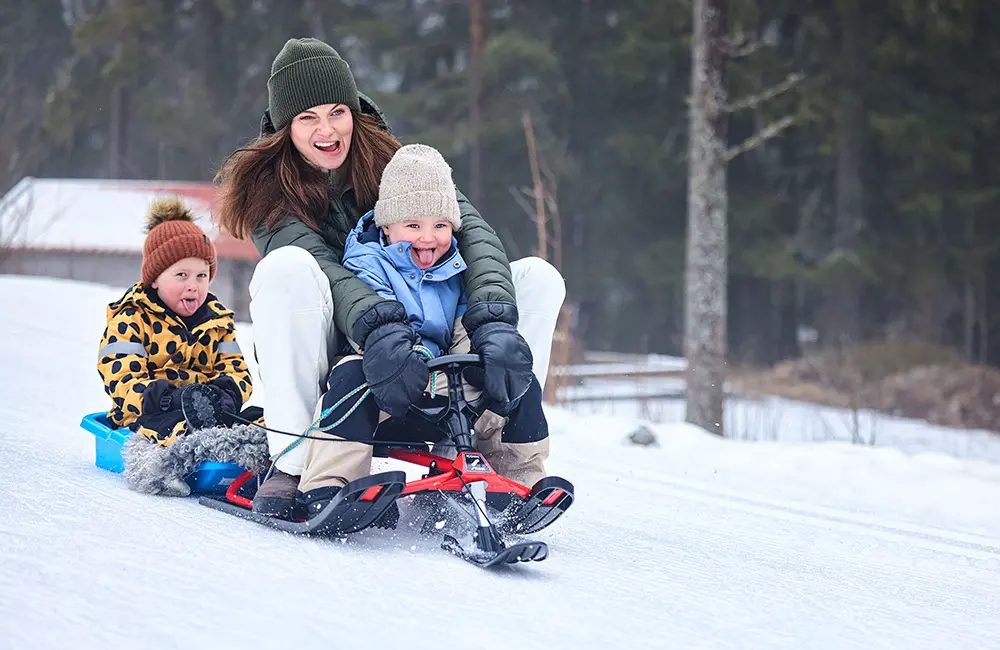 Kvinna i grön jacka och mössa sitter på en snowracer med ett barn framför sig och ett barn i pulka bakom. 