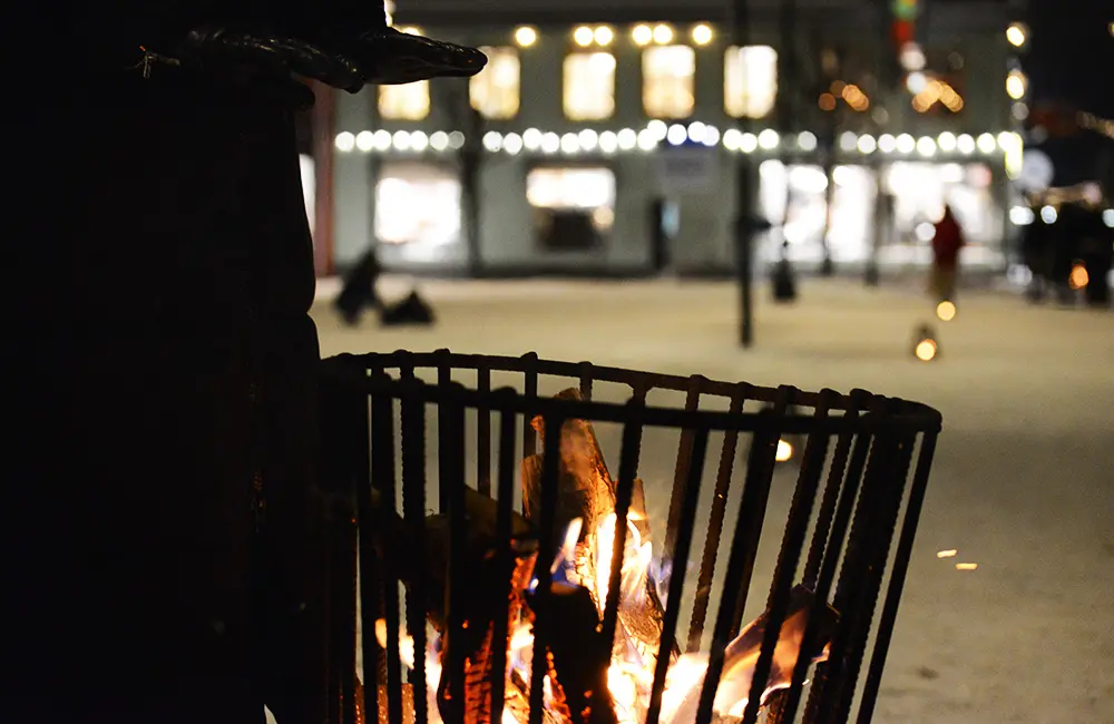 Snötäcktmark på torget med belysta butiker i bakgrunden. I förgrunden syns en eldkort och en person i svart jacka som värmer sig.
