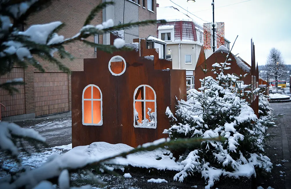 Pepparkakslikande hus i cortenplåt står på en gata. Runt är det snötäckta granar.