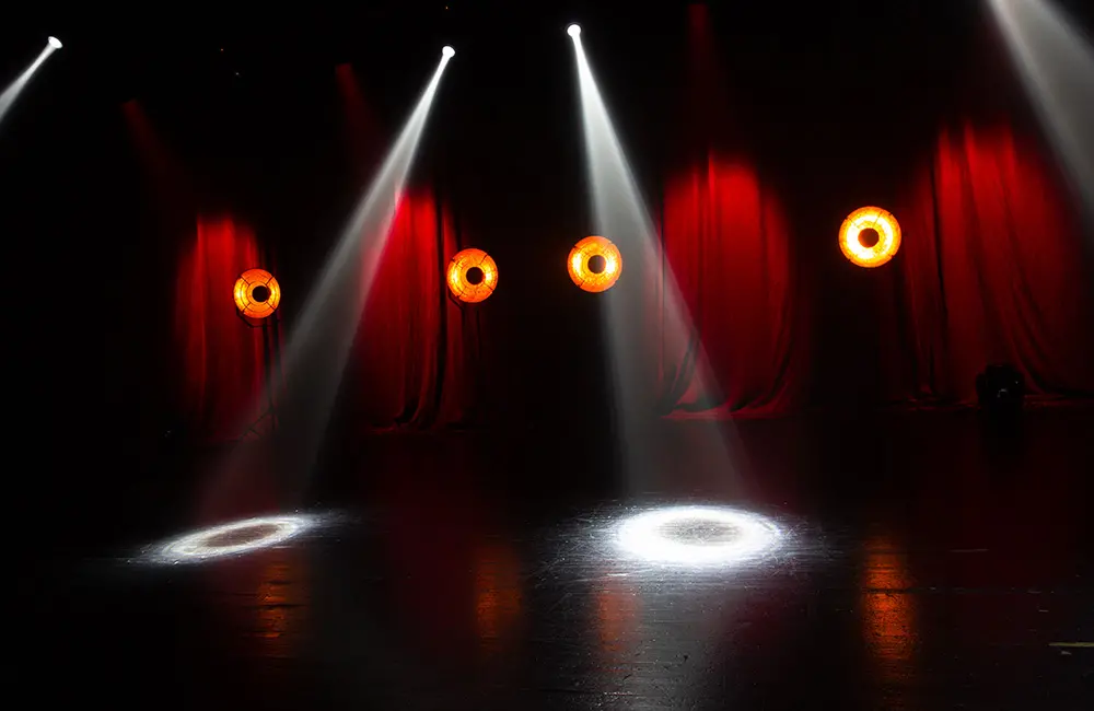 Scen med röda drapperier och strålkastare. Fyra gula lampor står på rad bakom scenen.