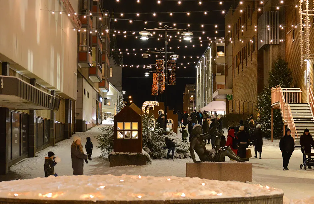 Gata med människor gåendes. Det är mörk på himlen och snö på marken. Marchaller är tända i en rund upphöjning och det är uppsatt julbelysning över gatan.