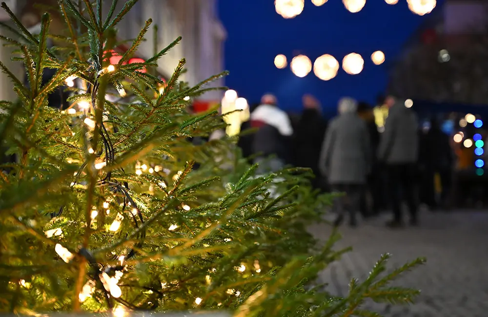 Gran med belysning till vänster i närbild, i bakgrunden syns suddigt en gågata med människor och julbelysning över gatan.