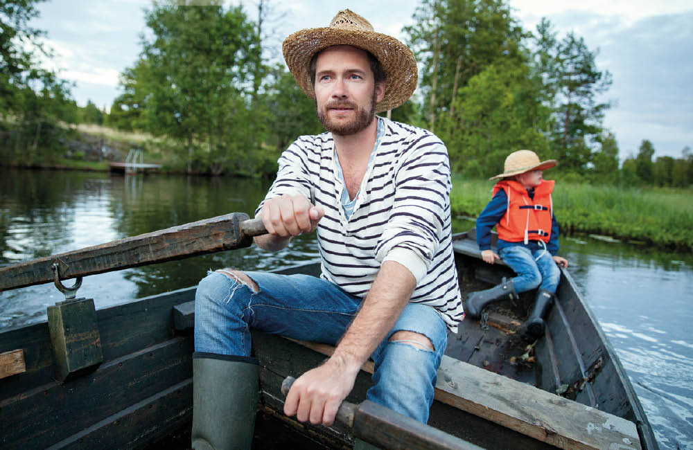 En man i stråhatt och randig tröja sitter i en roddbåt. I samma båt sitter även ett barn i hatt och flytväst.