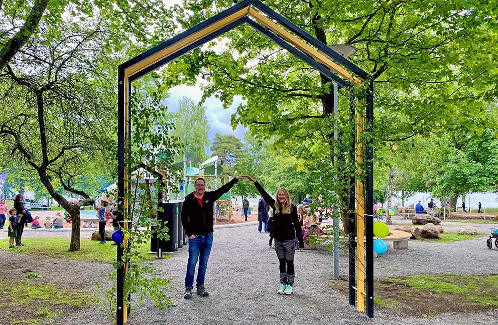 En man och en kvinna står i en park under en färgglad portal i metall. Deras fingertoppar möts i en båge över huvudet. för att bilda samma form som portalen.