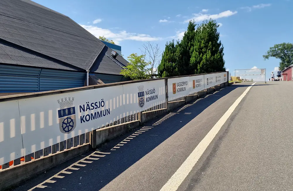 Staket med vita vepor vid en asfalterad väg. På veporrna är logotyper för Nässjö kommun och Vaggeryds kommun ihop med text. Bakom står ett blått hus med svart tak.