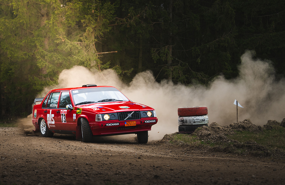 Röd rallybil kör i en kurva på en grusväg och gruset ryker  bakom. I bakgrunden är det grön skog och till höger en stapel med tre däck.