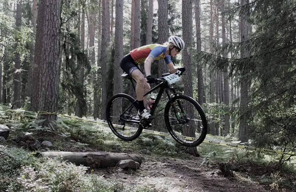 Kvinna i träningskläder och hjälm som cyklar bland träden i skogen.