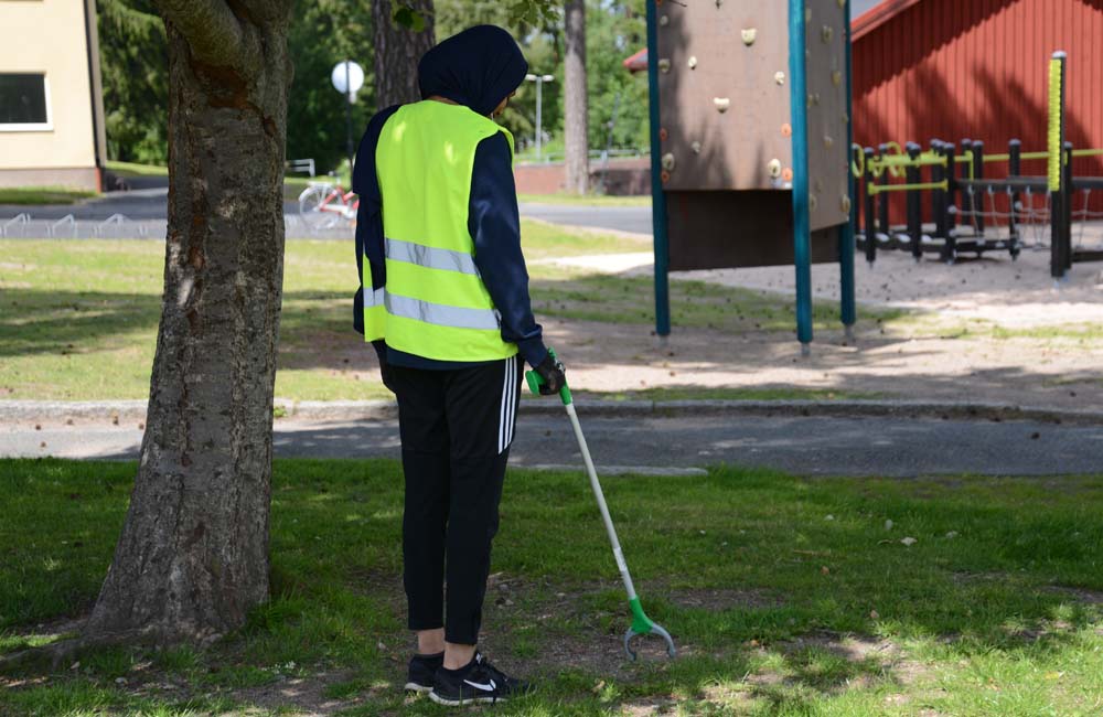 Feriepraktikant som plockar skräp vid lekplats.