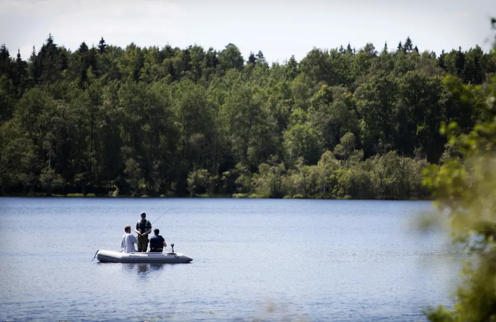 Tre personer i en båt på sjö, skog i bakgunden.