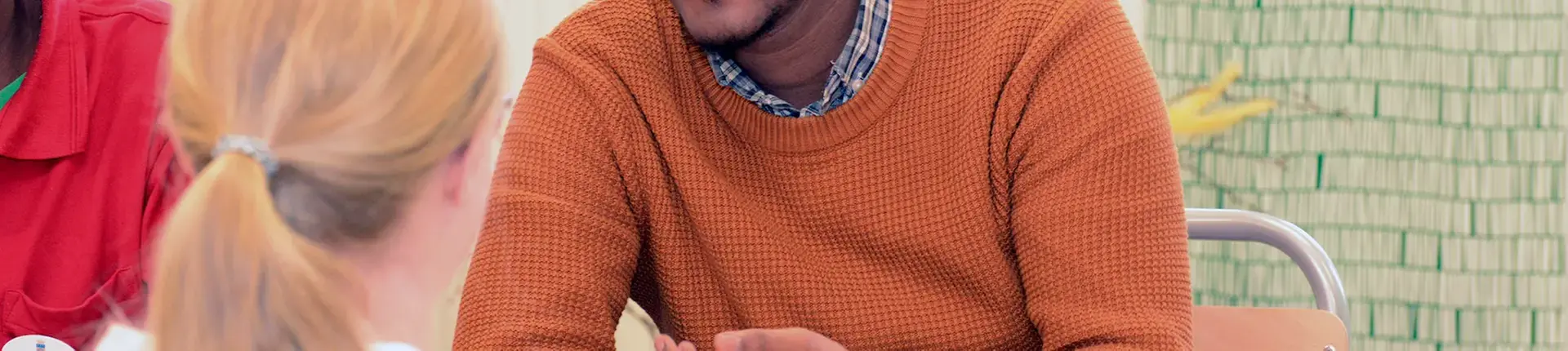 En tjej med ljus hästsvans sitter och pratar med en mörkhyad man i orange tröja. Man ser bara lite av deras ansikten.