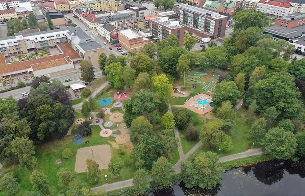 En flygbild över en grön lummig park i en stad. På marken syns en stor lekplats med olika, färger, lekytor och redskap.