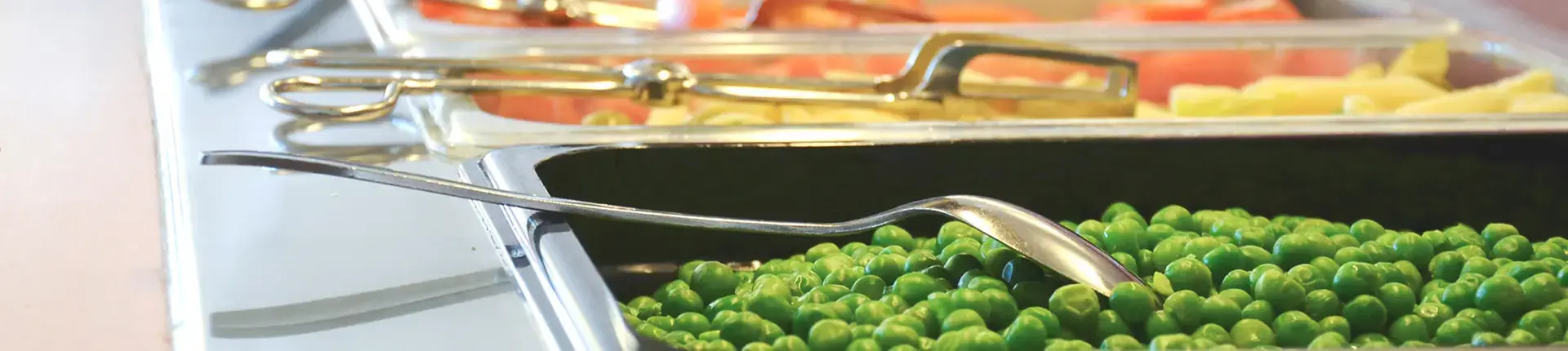 En närbild av en bufféservering med rostfria kantiner fyllda med olika livsmedel. I förgrunden syns en kantin med gröna ärtor och en metallsked placerad ovanpå. I bakgrunden finns fler kantiner med andra ingredienser, bland annat skivade grönsaker och potatis. Serveringsredskap i metall ligger ovanpå kantinerna. Ljuset är klart och reflekteras i de blanka ytorna.