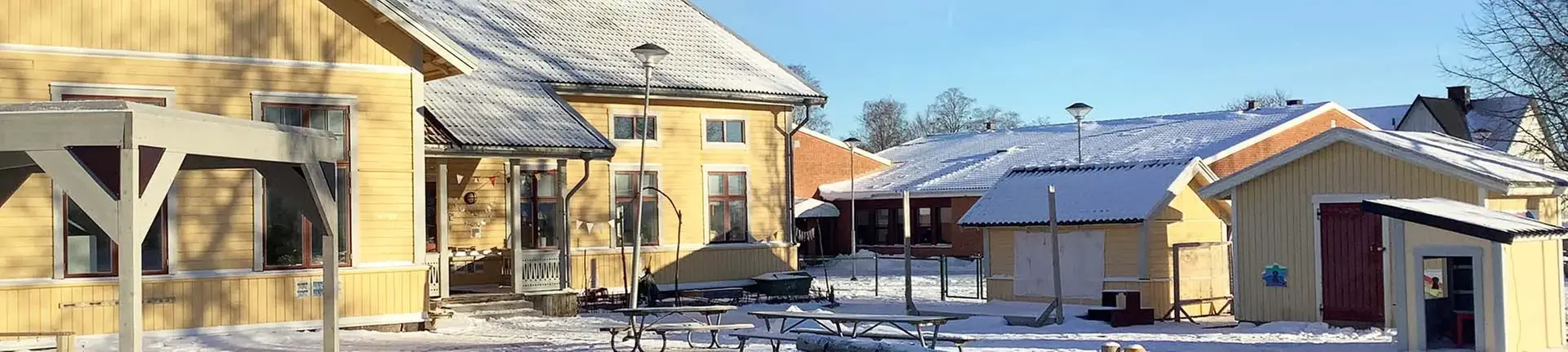 Bodafors västra förskola. Gula träbyggnader samlade omkring en skolgård. Bilden är tagen på vintern.