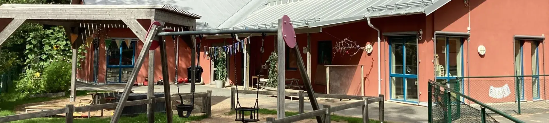 En terrakottafärgad byggnad med omkringliggande aktivitetsyta innehållande sand och gungor.