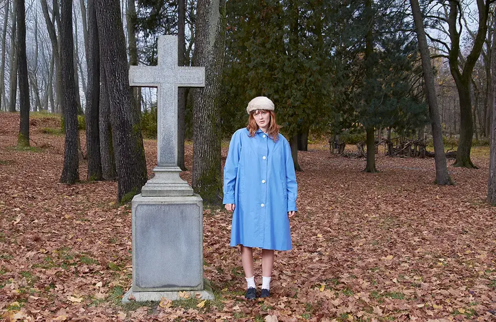 Kvinna i blå lång skjorta står bland höstlöv bredvid ett kors i cement.
