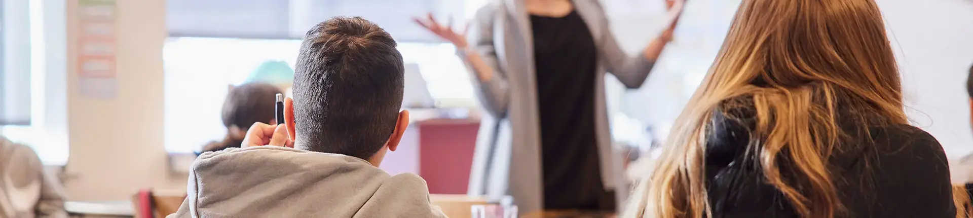 Två elever som lyssnar och räcker upp handen. Framför eleverna står en lärare.