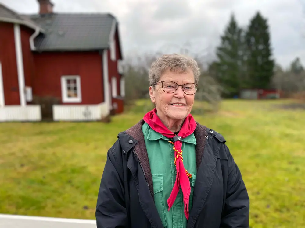 En äldre kvinna i grön scoutskjorta och röd scarf står utomhus och ler in i kameran. I bakgrunden syns ett rött hus och en gräsmatta.
